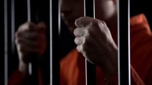 Hands of a person in an orange jumpsuit holding jail cell bars