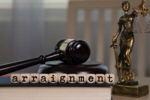 Word ARRAIGNMENT composed of wooden dice. Wooden gavel and statue of Themis in the background. Learn what happens during an arraignment for murder in Arizona.