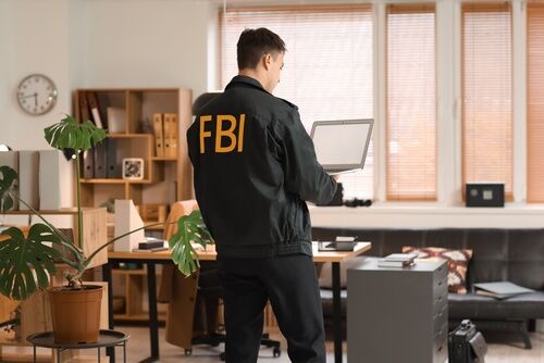 FBI jacket lettering visible as agent stands holding laptop in office
